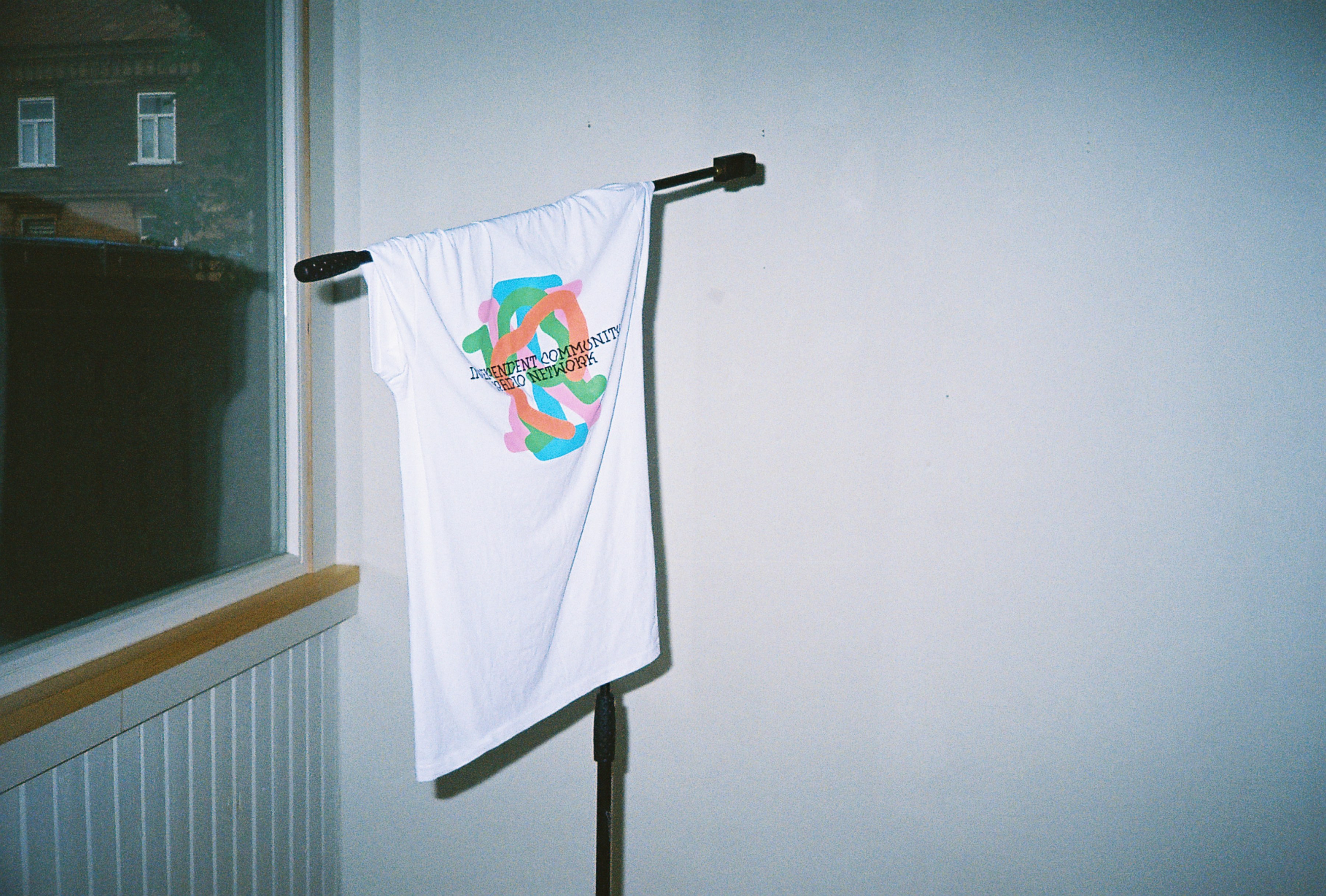 A color photo of a t-shirt hanging from a microphone stand. The t-shirt is white and has a print that says: Independent Community Radio Network.